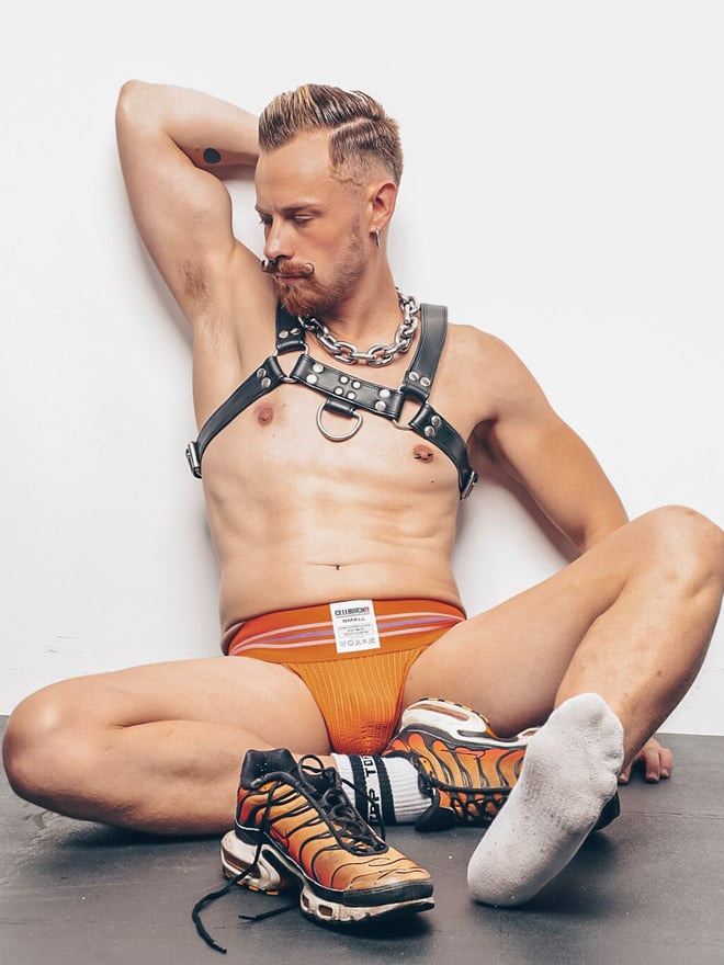 Man with short blonde hair and tattoos posing shirtless in leather harness and orange underwear against a plain wall, with sneakers nearby.