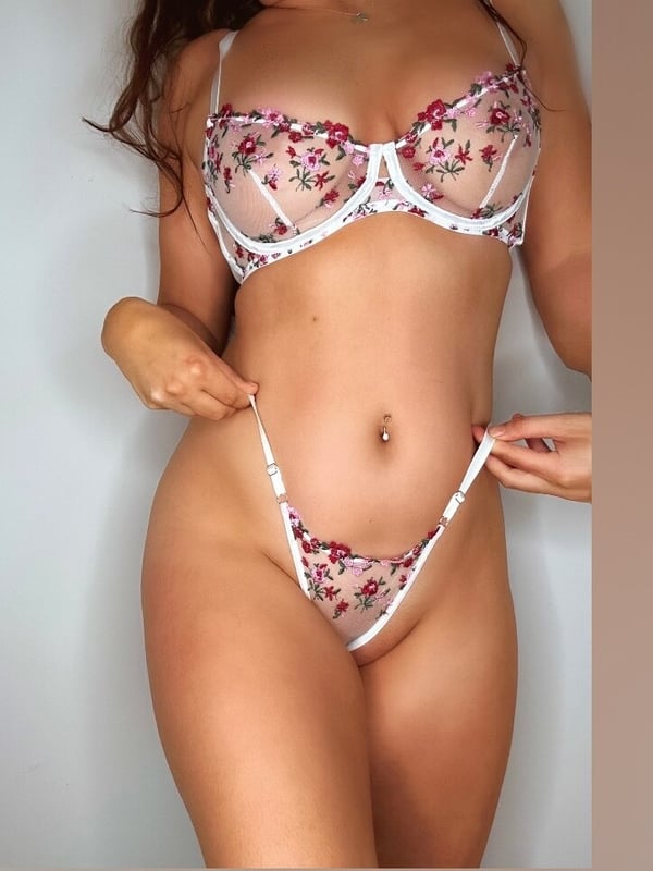 A person posing in sheer floral lingerie, showing a navel piercing and standing confidently.