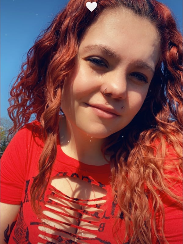 A young woman with curly red hair in pigtails smiling outdoors, wearing a red top with cutouts under a blue sky.