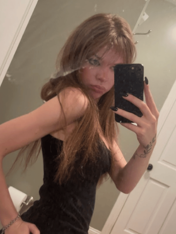 A person taking a mirror selfie wearing a black dress, with long brown hair and visible tattoos on the arm.