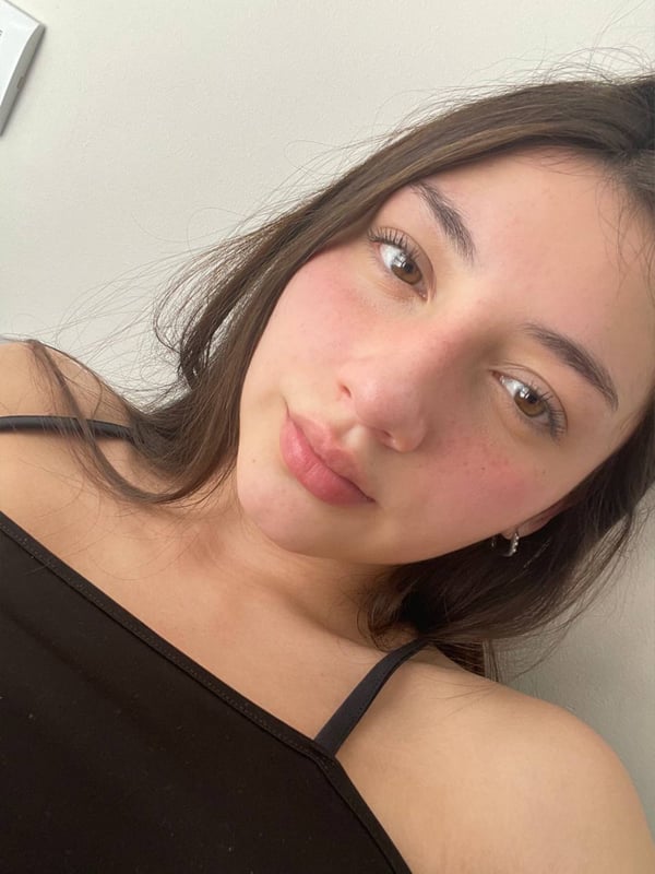 Close-up selfie of a young woman with long brown hair and flushed cheeks, wearing a black tank top, looking at the camera in an indoor setting.