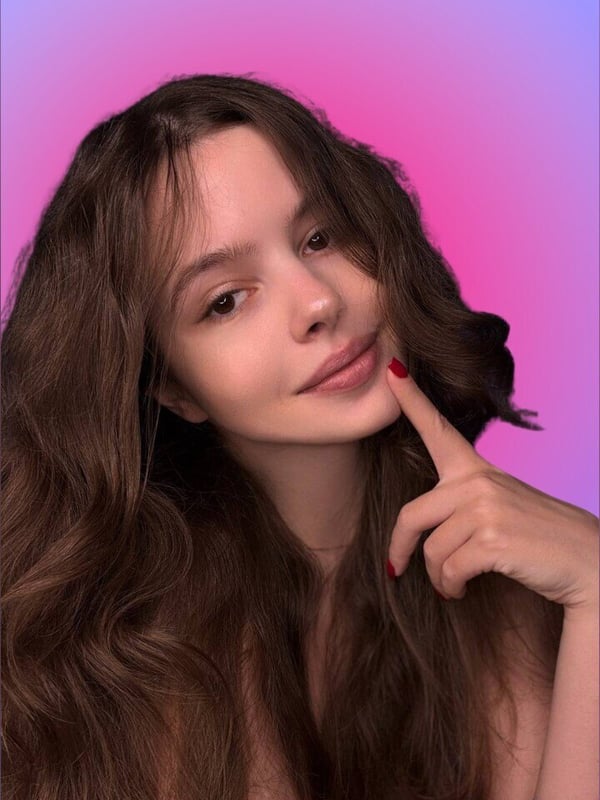 A young woman with long brown hair and red nails making a playful gesture with her finger to her lips, smiling against a vibrant purple and pink gradient background.