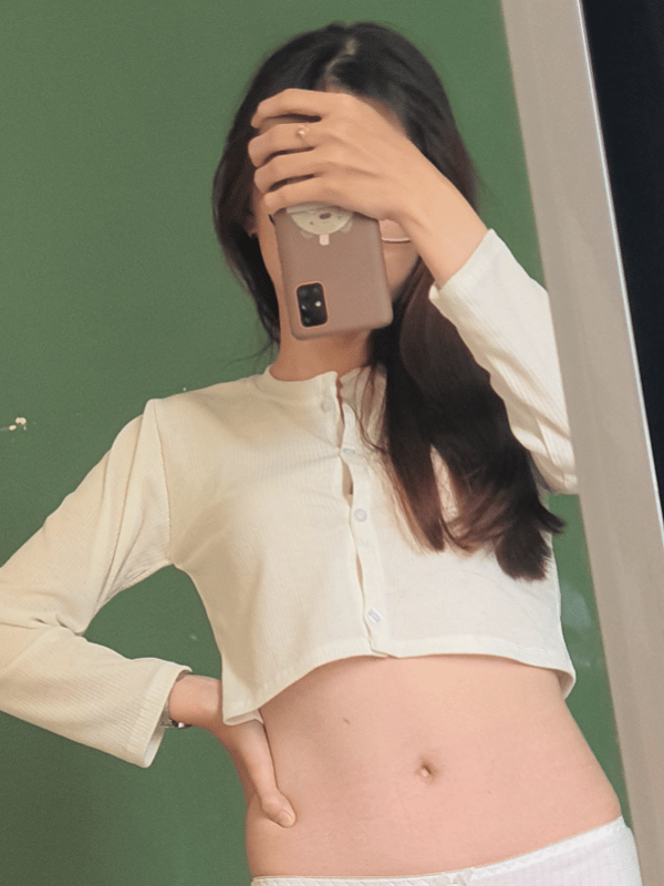 Young Asian woman in a white cropped top and white pants posing in front of a green wall while holding her phone for a selfie.