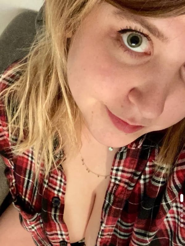 A young woman with blonde hair smiling at the camera, wearing a plaid shirt while seated indoors.