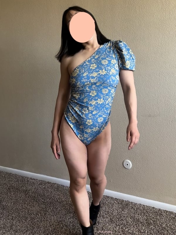 Woman standing in a floral-patterned one-shoulder bodysuit and black boots against a neutral indoor background.