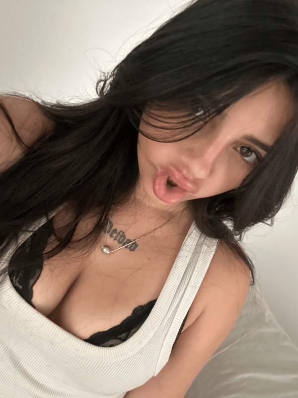 A young woman with long dark hair taking a selfie indoors while wearing a white top.