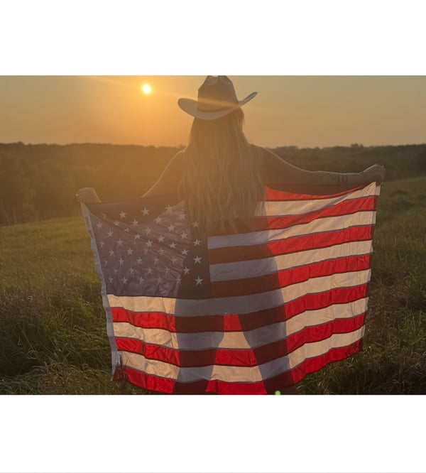 A woman in a cowboy hat holding an American flag, standing with her back to the sunset over a grassy field.