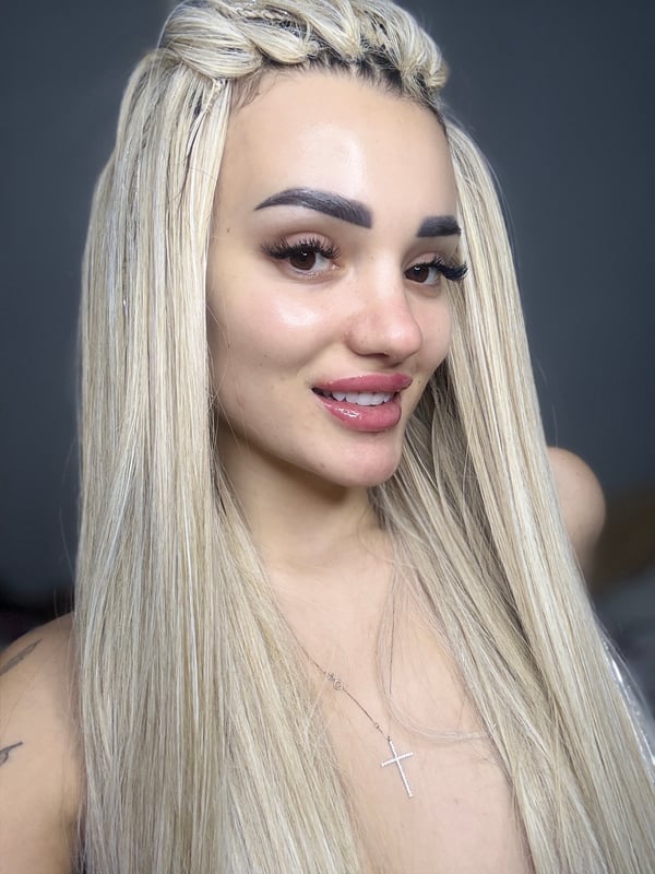 Close-up portrait of a woman with long blonde braids, bold makeup, and a subtle smile.
