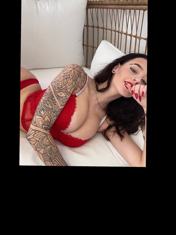 A woman with long dark hair and tattoos poses reclined on a bed with white pillows and a wicker headboard, wearing red lingerie.