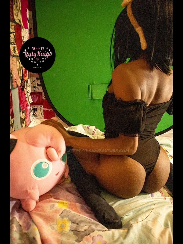 A woman in cosplay attire posing on a bed next to a plush toy in a colorful room with gaming decor.