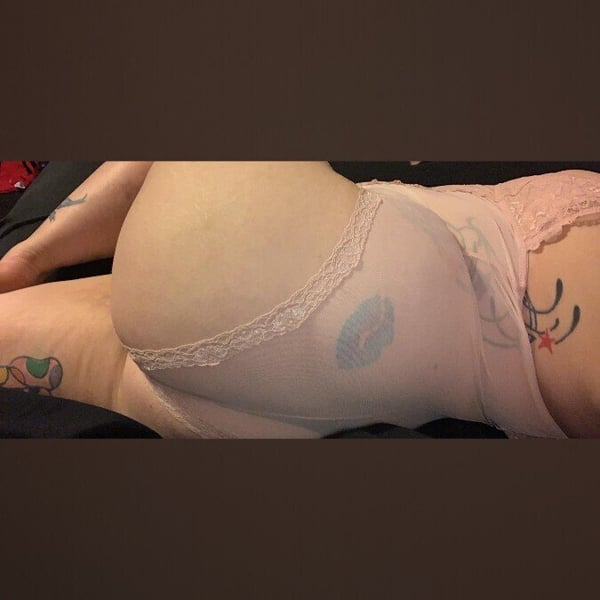 Person reclining on a bed in lingerie, showcasing tattoos on legs and hips against a dark background.