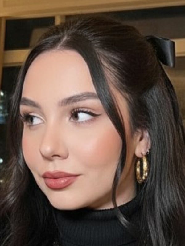 A woman with dark hair and gold hoop earrings smiling elegantly in a black turtleneck against a warm indoor background.