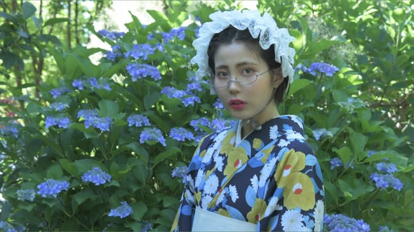 A woman wearing a traditional floral kimono and white headpiece poses gracefully in front of lush green hydrangea bushes.