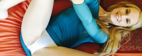 A smiling woman with long blonde hair reclining on a red couch, wearing a blue top and white shorts.