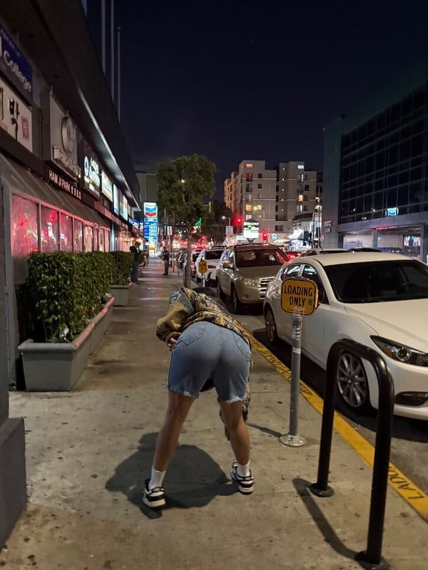 Person bending over near a parking sign on a vibrant urban street at night with neon lights and cars in the background.