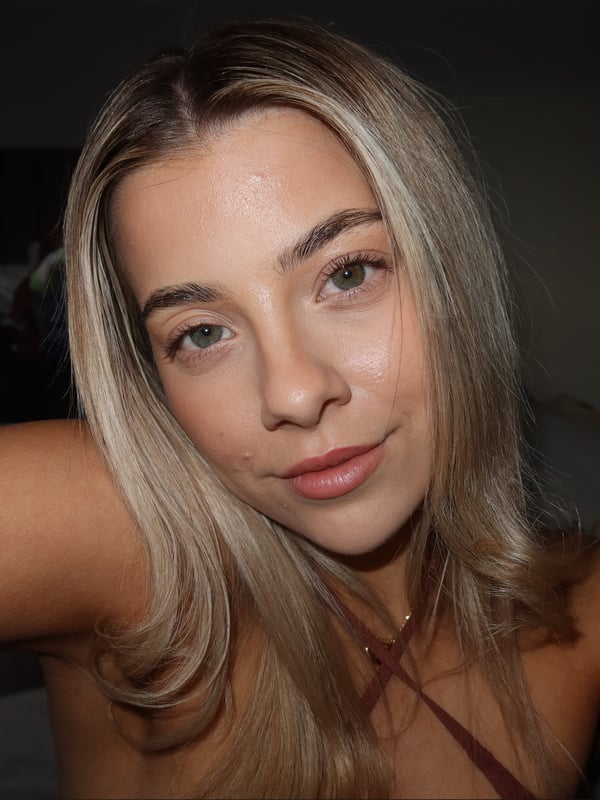 Close-up selfie of a young woman with blonde hair smiling at the camera in a dimly lit room.