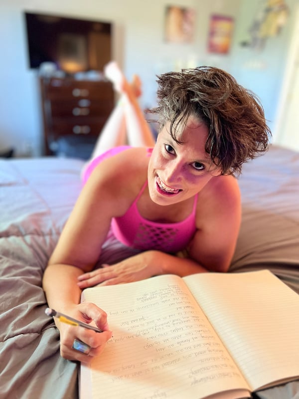 A woman with curly brown hair lounging on a bed in a pink top, smiling while writing in a notebook in a cozy bedroom with art on the walls.