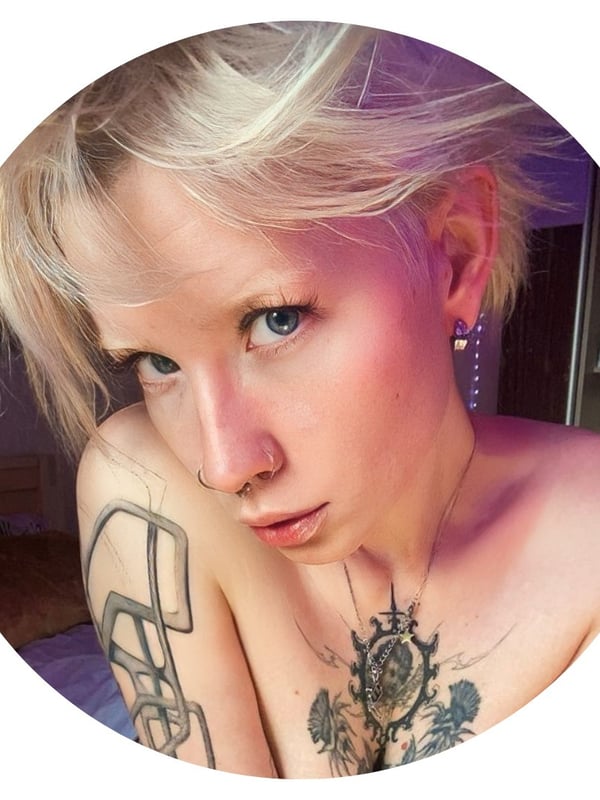 Close-up portrait of a young blonde woman with blue eyes, tattoos on her shoulder and chest, and earrings, smiling softly indoors.