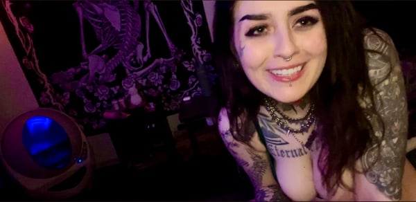 A woman with tattoos and piercings smiling at the camera in a dimly lit room with gothic decor and purple lighting.