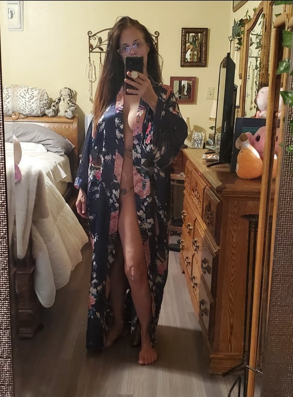 Woman standing in a bedroom wearing a floral robe, holding a phone for a selfie, with home decor in the background.