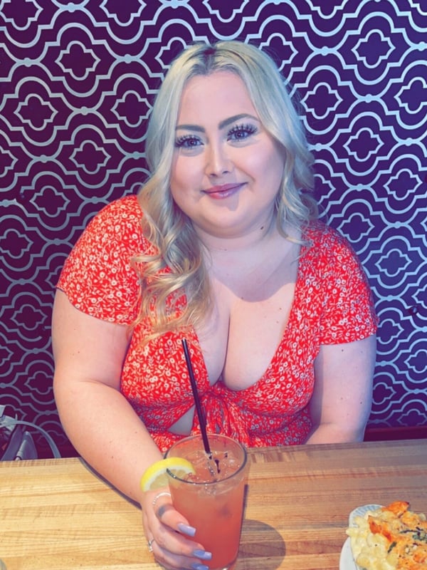 A smiling woman with blonde hair sitting at a table, wearing a red floral dress, holding a pink drink with a lemon garnish.