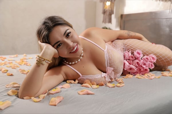 A woman with a warm smile reclines on a bed surrounded by rose petals, wearing elegant lingerie and jewelry in a cozy bedroom setting.