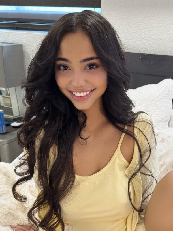 Young woman with long wavy dark hair smiling happily in a bedroom, wearing a yellow off-shoulder top against white pillows.