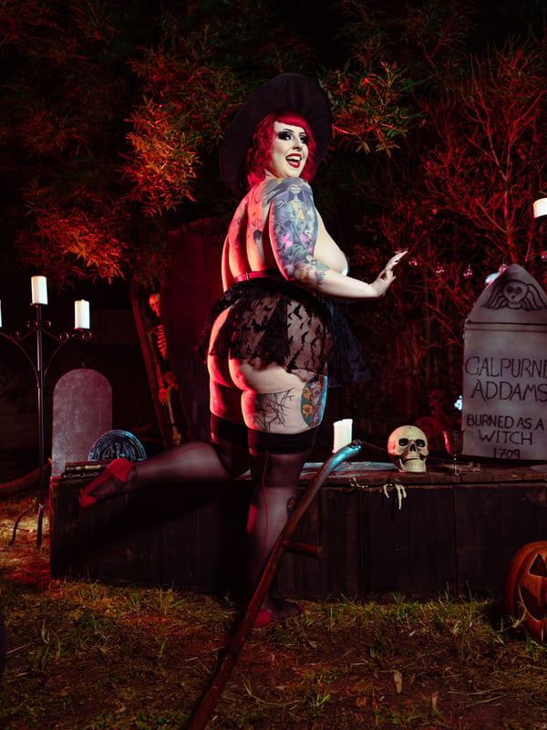 A plus-size woman in a black witch costume with red hair and tattoos stands confidently near a gravestone in a spooky outdoor Halloween setting with candles and pumpkins.