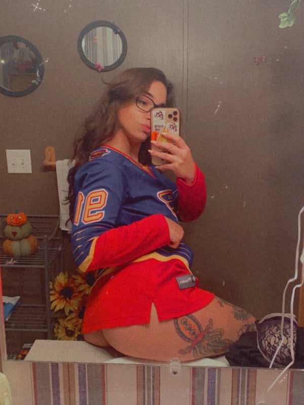 A woman with long dark hair takes a mirror selfie in a bathroom, dressed in a blue sports jersey and red shorts, with tattoos visible on her leg.