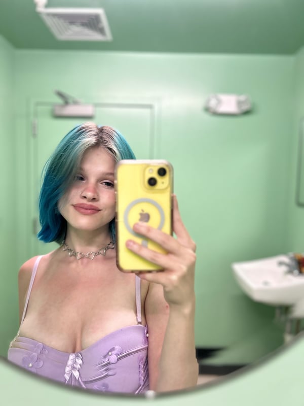 Young woman with colorful dyed hair posing in a black top against a rainbow flag background in a bedroom setting.