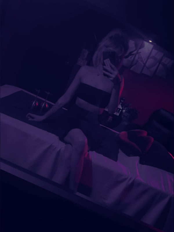 A woman with blonde hair sits on a purple surface in a low-lit room, smiling while covering part of her face with her hand.