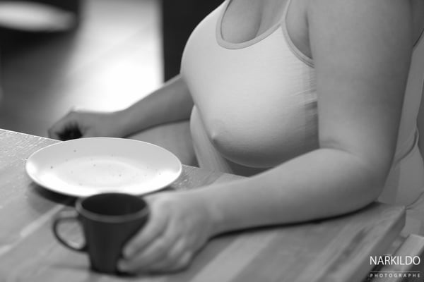 Woman in a white tank top sitting at a table, holding a cup and with a plate in front of her, in black and white photography.