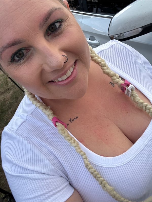 Smiling woman with blonde braids and nose piercing, posing in a white top next to a car.