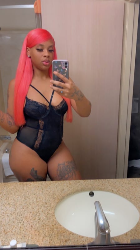 A woman with long pink hair posing in a black lace outfit in front of a bathroom mirror, holding her phone for a selfie.
