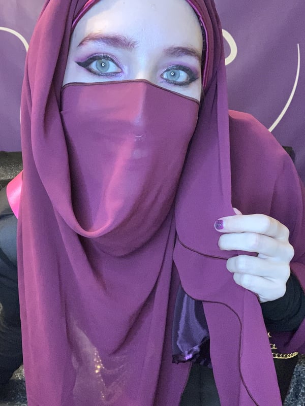A woman wearing a purple hijab and niqab, posing on a bed with a purple backdrop.