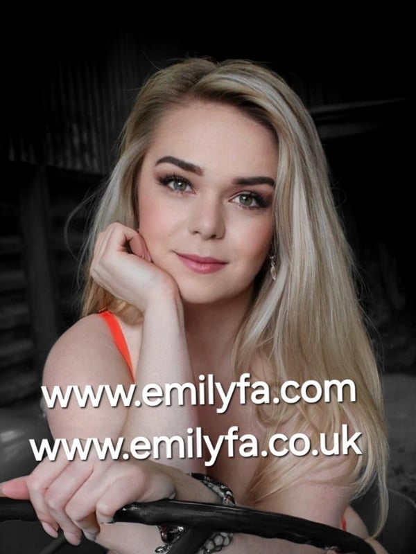 Blonde woman with long hair smiling, leaning on a steering wheel in an orange top against a dark background.