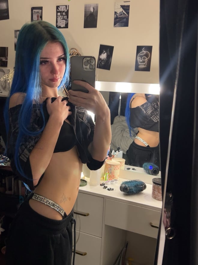 A young woman with long blue hair posing for a mirror selfie in a checked top.