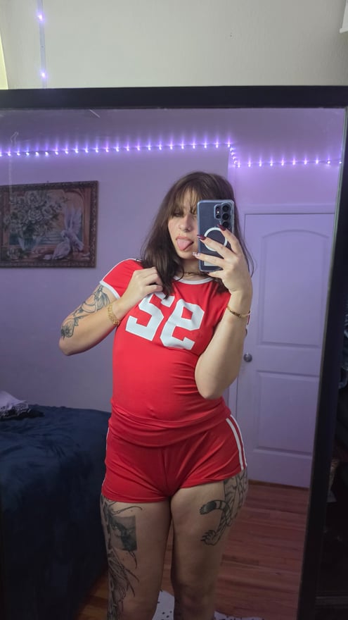 A tattooed woman poses in black lingerie while taking a mirror photo in a warmly lit room.