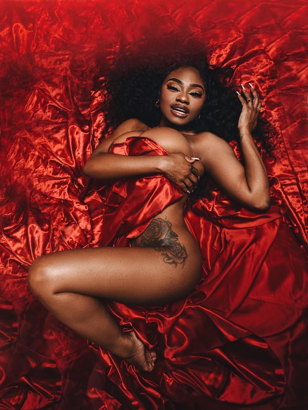 A woman with curly hair lying on red silk sheets, holding fabric against her chest in a dramatic pose.