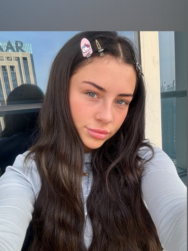 Young woman with long dark hair and hair clips, posing with a subtle expression in front of a window with a city building view.