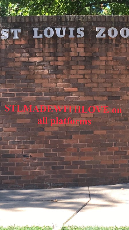 Brick wall at the Saint Louis Zoo entrance with promotional text for STLMADEWITHLove on all platforms against a green outdoor background.