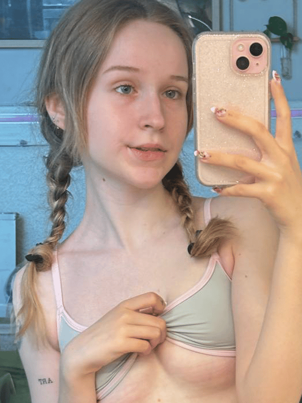 Young woman with blonde pigtails taking a mirror selfie in a bedroom, holding her phone and smiling softly.