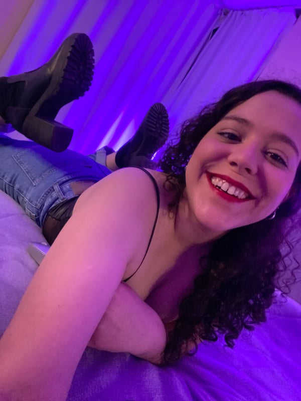 Young woman with curly dark hair smiling at the camera, lying on a bed in a dimly lit purple room while wearing jeans and a tank top.