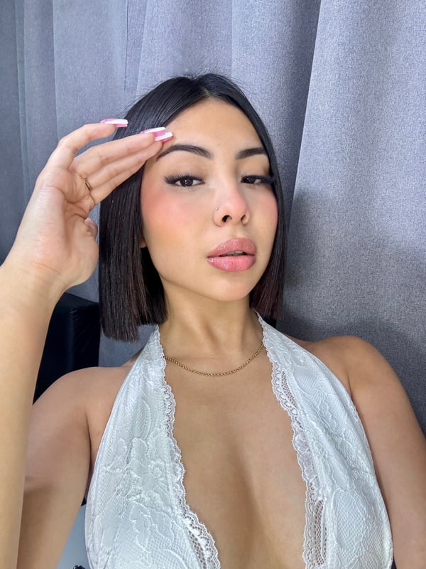 A person with dark hair wearing a white lace top, posing with one hand near their face against a gray curtain.