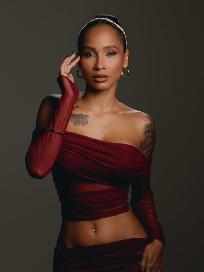 A woman poses in a burgundy outfit against a dark studio background, wearing hoop earrings and visible tattoos.