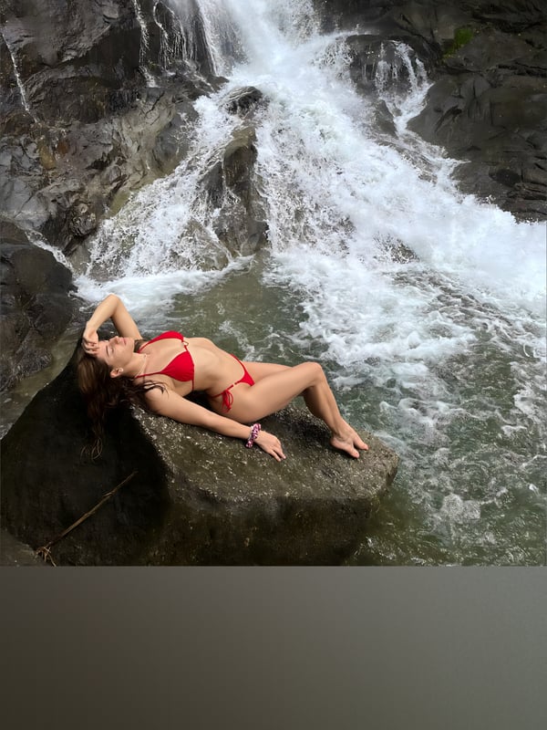 Woman in a red bikini lounging on a rock beside a cascading waterfall in a natural rocky setting.