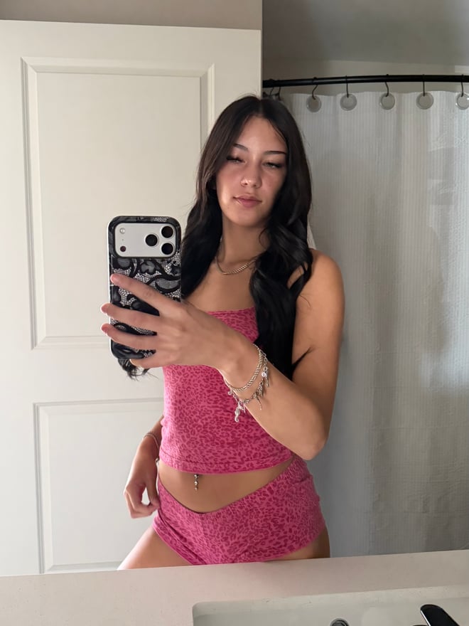 Young woman with long dark hair posing in a zebra-print bodysuit and fluffy boots, taking a mirror selfie in a bedroom with warm lighting.