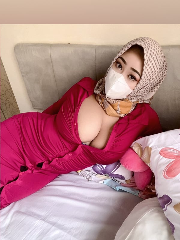 A woman wearing a patterned headscarf and face mask reclines on a bed in a pink outfit.