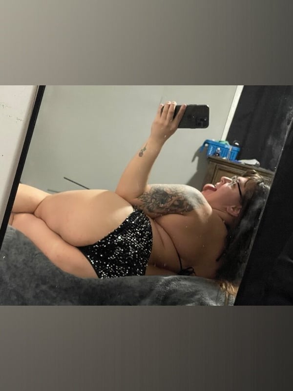 A woman taking a mirror selfie while lying on a bed, smiling widely with tattoos visible on her arm, dressed in sparkling black lingerie.