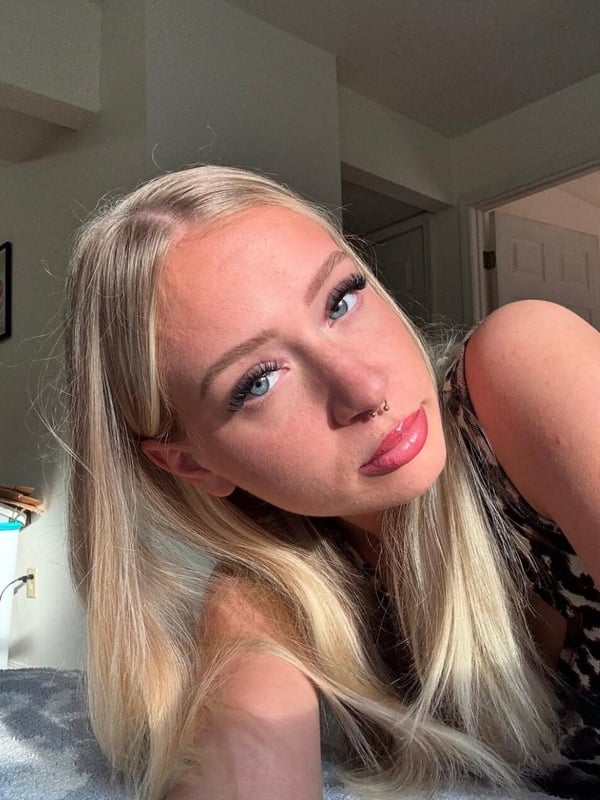 A close-up selfie of a young woman with long blonde hair and blue eyes, wearing pink lipstick.
